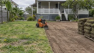 Queensland backyard half weedy old lawn, half stripped to bare soil, with an orange turf cutter and stacked turf on pallets