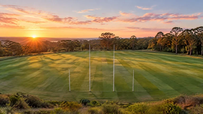 Striped Australian football oval at sunrise, white goalposts in front of eucalypt bushland